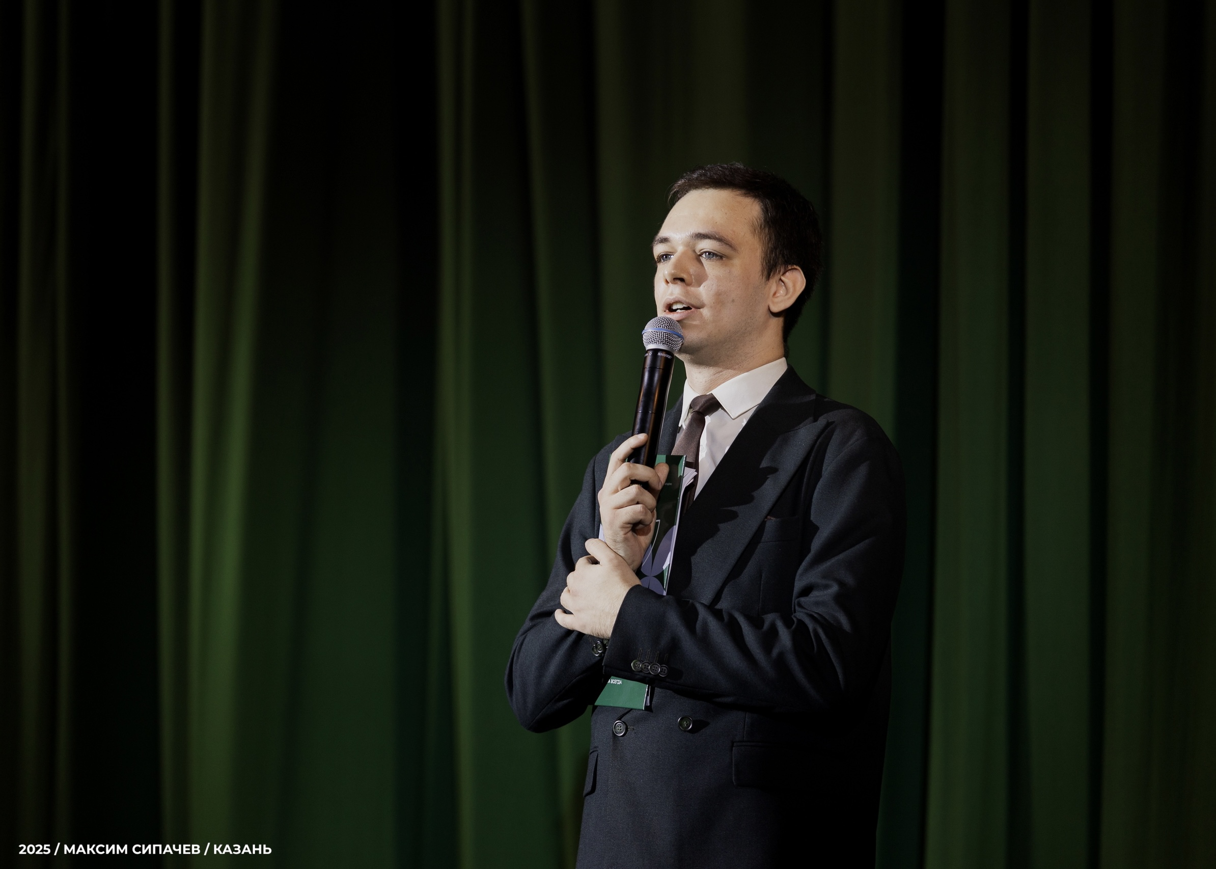 Руслан Балдин выступает с микрофоном на фоне зелёного бархата